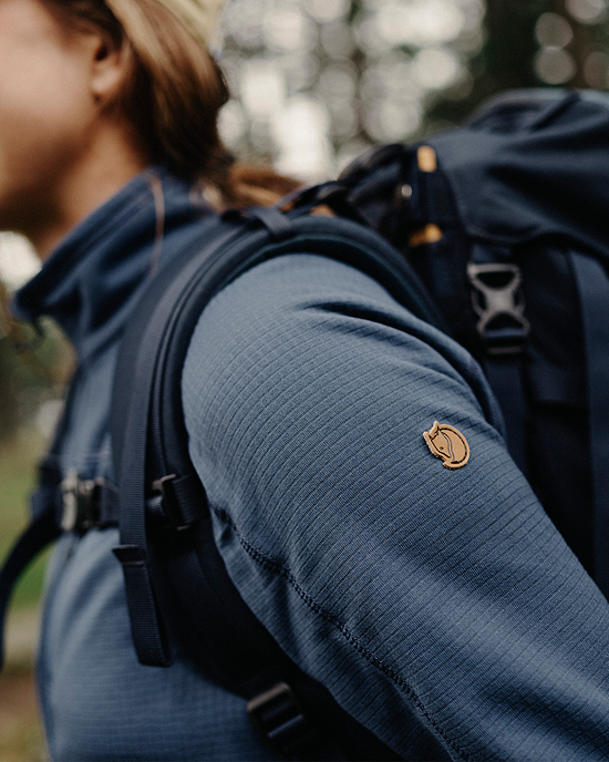A close‑up of a person's shoulder wearing a blue textured jacket with a logo, carrying a backpack in a natural setting.