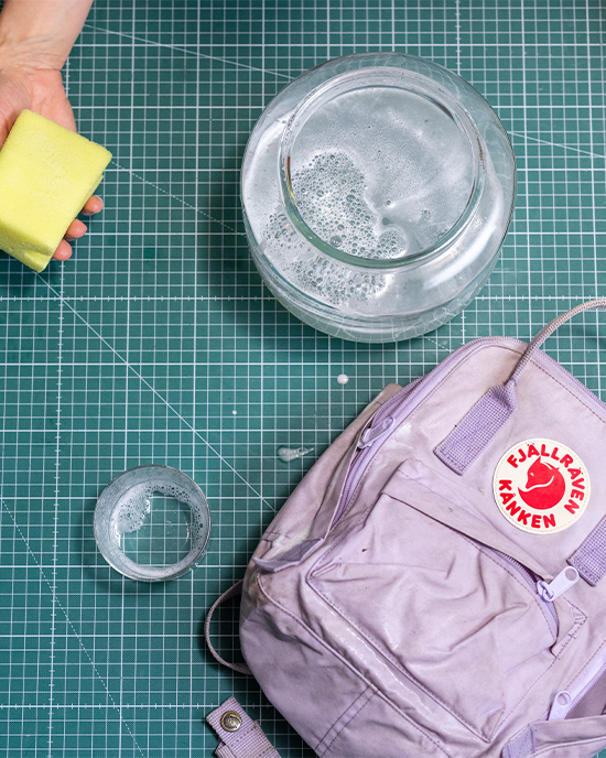 A yellow sponge is held above a bowl of soapy water, next to a light purple backpack on a green cutting mat.